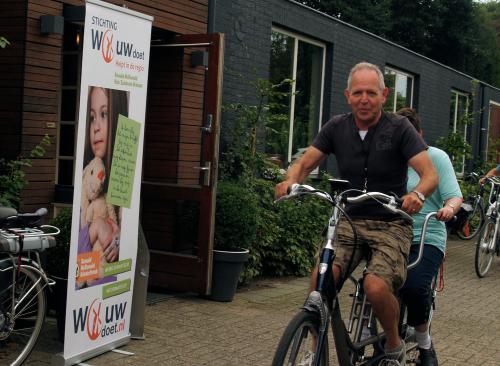 Johan rijdt met Esmeralda op de tandem langs een banner van ''Wouw Doet''